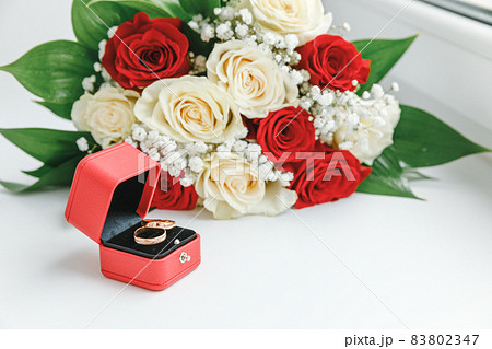 Gold wedding rings and flowers bouquet in white red colors lying on white background. Declaration of love, spring. Wedding card, Valentine's Day greeting. Wedding rings. Wedding day details. 83802347