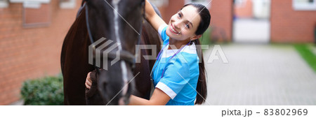 Smiling female veterinarian hugs black horse near stables Smiling female veterinarian hugs black horse near stables 83802969