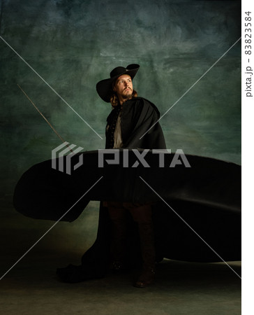 Full-length portrait of genteel brutal man, medeival pirate in hat and cloak with sword isolated over dark background. Full-length portrait of genteel brutal man, medeival pirate in hat and cloak with sword isolated over dark background. 83823584