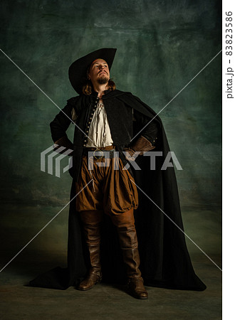 Full-length portrait of genteel brutal man, medeival pirate in hat and cloak with sword isolated over dark background. Full-length portrait of genteel brutal man, medeival pirate in hat and cloak with sword isolated over dark background. 83823586