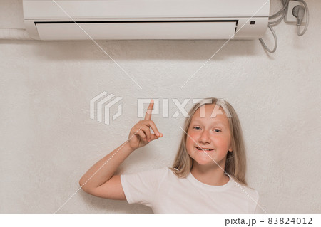 Cute teenage girl blonde European appearance points a hand finger at the air conditioner on the wall in the room 83824012
