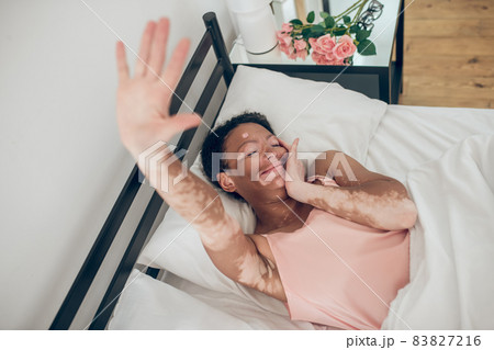 A young dark-skinned woman lying in bed, stretching hand and smiling 83827216