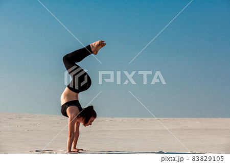 Young woman practicing handstand on beach with white sand and bright blue sky 83829105