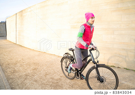 Young active woman in warm wear riding bike outdoors near wall on streets 83843034