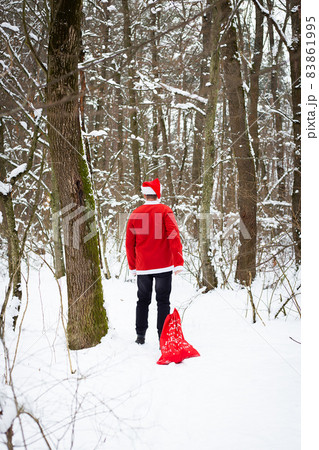 A traditional Santa Claus in a hat and a bag of gifts walks through the forest carrying gifts. Back view. A traditional Santa Claus in a hat and a bag of gifts walks through the forest carrying gifts. Back view. 83861995