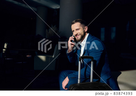 Handsome European businessman talks on cell phone sitting on an arm chair in hotel lounge and smiles looking aside. Successful investment advisor with a luggage talks on mobile phone in hotel lobby Handsome European businessman talks on cell phone sitting on an arm chair in hotel lounge and smiles looking aside. Successful investment advisor with a luggage talks on mobile phone in hotel lobby 83874393
