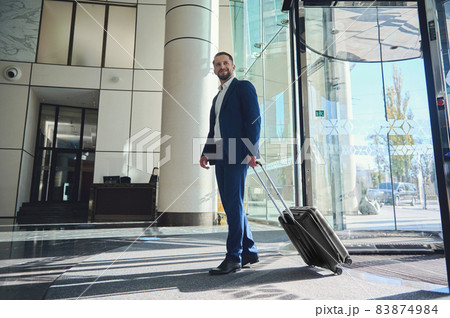A full-length handsome businessman developer entrepreneur in a business suit with a suitcase stands at the entrance of a modern luxury building, walking to the lobby during his business trip A full-length handsome businessman developer entrepreneur in a business suit with a suitcase stands at the entrance of a modern luxury building, walking to the lobby during his business trip 83874984