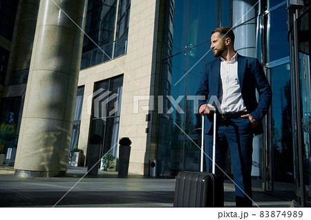 Full-length portrait of confident businessman in casual business suit standing with black suitcase at the glass entrance of the hotel, looking confidently to the side, holding his hand in his pocket Full-length portrait of confident businessman in casual business suit standing with black suitcase at the glass entrance of the hotel, looking confidently to the side, holding his hand in his pocket 83874989
