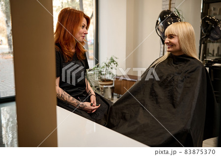 Fashionable pretty red-haired woman in black professional uniform, hair stylist with stylish body tattoos on arms talking with a hair salon customer sitting in barber chair in front of a mirror Fashionable pretty red-haired woman in black professional uniform, hair stylist with stylish body tattoos on arms talking with a hair salon customer sitting in barber chair in front of a mirror 83875370