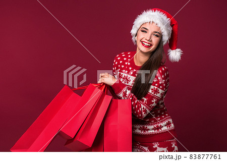 Young pretty woman or girl with long beautiful hair in Santa hat and New Year's dress holding red package on red studio background Young pretty woman or girl with long beautiful hair in Santa hat and New Year's dress holding red package on red studio background 83877761