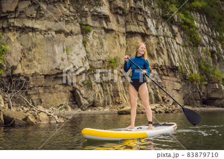 Young women Having Fun Stand Up Paddling in the sea. SUP. Red hair girl Training on Paddle Board near the rocks Young women Having Fun Stand Up Paddling in the sea. SUP. Red hair girl Training on Paddle Board near the rocks 83879710