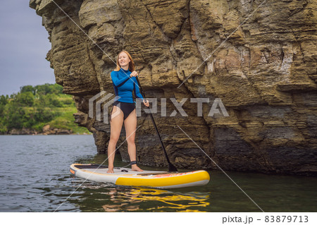 Young women Having Fun Stand Up Paddling in the sea. SUP. Red hair girl Training on Paddle Board near the rocks Young women Having Fun Stand Up Paddling in the sea. SUP. Red hair girl Training on Paddle Board near the rocks 83879713