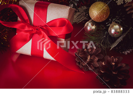 New Year's gift with a red ribbon in craft brown paper. Christmas composition of cones, tinsel and gifts on a red background. 83888333