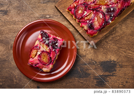 Piece of plum tart on ceramic plate, next to pie on wooden board 83890373