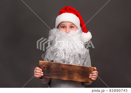 a boy 8-10 years old in a hat and with a Santa beard. He holds a wooden sign in his hands for advertising and Christmas sale. on a gray background. The concept of New Year sales. copy space a boy 8-10 years old in a hat and with a Santa beard. He holds a wooden sign in his hands for advertising and Christmas sale. on a gray background. The concept of New Year sales. copy space 83890745