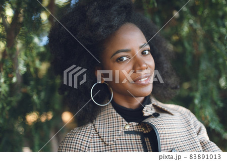 Beautiful African American young woman with afro and large hoop earrings in a stylish coat in a spring or autumn park, smiling. Selective focus, copy space Beautiful African American young woman with afro and large hoop earrings in a stylish coat in a spring or autumn park, smiling. Selective focus, copy space 83891013