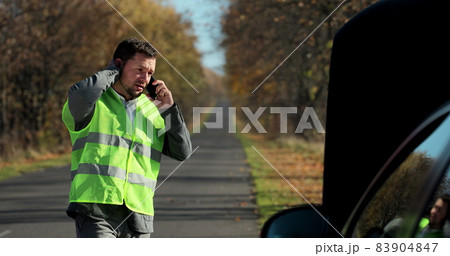 Portrait of young man in a safety vest standing in front of his broken car, looking at car engine and talking on cell phone. Helpless man need help and assistance. Portrait of young man in a safety vest standing in front of his broken car, looking at car engine and talking on cell phone. Helpless man need help and assistance. 83904847