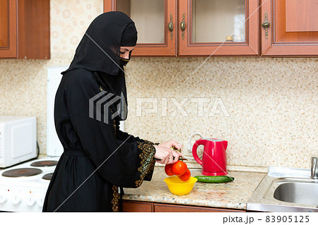A Muslim woman in national clothes cooks lunch in the kitchen slicing tomatoes A Muslim woman in national clothes cooks lunch in the kitchen slicing tomatoes 83905125
