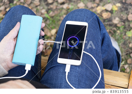 The phone is charged from a power bank in hands on knees in jeans The phone is charged from a power bank in hands on knees in jeans 83913555