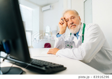 Portrait of gray-haired mature adult male doctor wearing white uniform with stethoscope sitting at desk with computer in hospital, looking at camera. 83916296