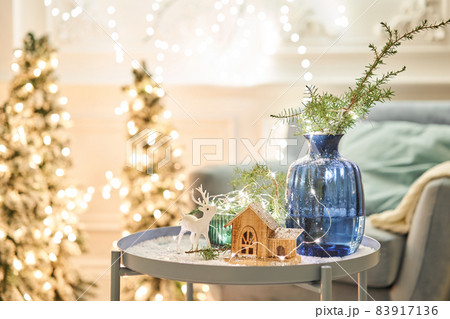 Decorated table in the Christmas interior. Christmas tree. Close-up a small house, Cones, garland, and other Christmas tree toys. light from the garlands 83917136