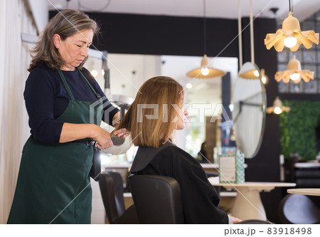 Aged woman hair stylist making haircut to young female client 83918498