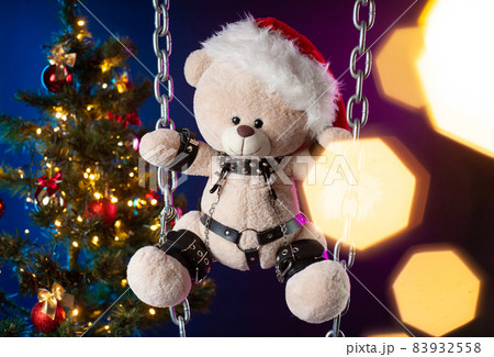 A teddy bear in a Santa Claus hat is a Christmas gift for BDSM games on the background of a Christmas tree A teddy bear in a Santa Claus hat is a Christmas gift for BDSM games on the background of a Christmas tree 83932558