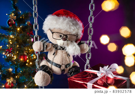 A teddy bear in a Santa Claus hat is a Christmas gift for BDSM games on the background of a Christmas tree 83932578