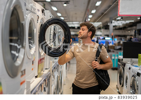 Man in casual clothes opening door and looking inside contemporary washing machine while choosing appliances in mall 83939025