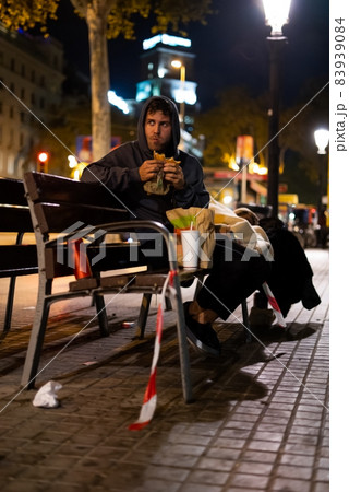 Homeless guy in hoodie chewing burger and looking away while sitting on bench on illuminated street at night 83939084