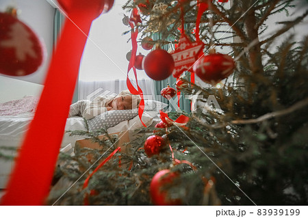 Photo of a boy in pajamas on a bed next to a Christmas tree decorated with red toys 83939199