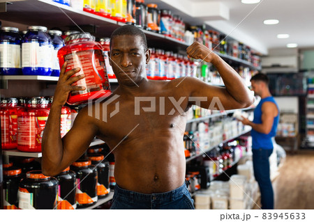 Bare-chested athletic African standing with cans of sports nutrition in store, demonstrating muscles Bare-chested athletic African standing with cans of sports nutrition in store, demonstrating muscles 83945633
