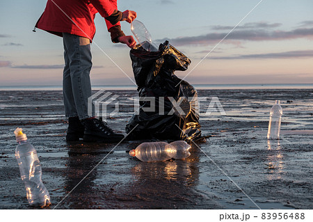 A woman cleans the bank of plastic bottles and puts the trash in a garbage bag. Environmental pollution by plastic. Recyclable packaging. 83956488