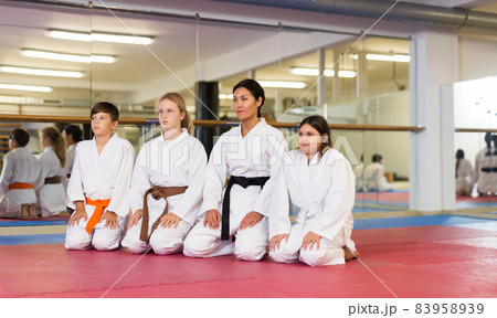 Group of children dressed in kimonos sitting on floor at gym with female trainer 83958939