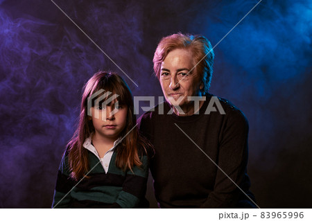 serious and angry expression of grandmother and granddaughter looking at the camera with smoke and colored lights. portrait in studio serious and angry expression of grandmother and granddaughter looking at the camera with smoke and colored lights. portrait in studio 83965996