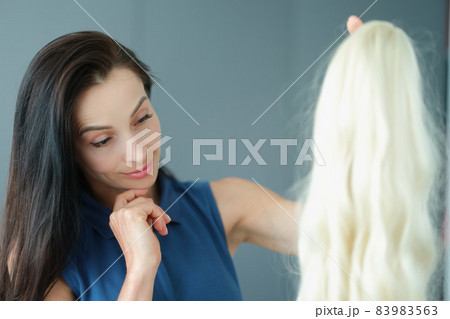 Brunette woman holding blonde wig with long hair and thinking to wear it 83983563