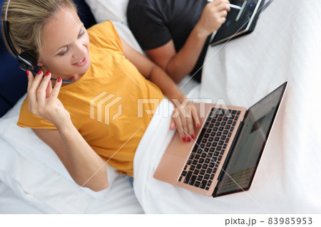 Young woman in headphones with microphone works on laptop while lying in bed Young woman in headphones with microphone works on laptop while lying in bed 83985953