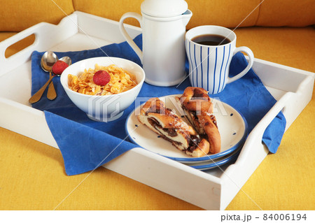 Rich continental breakfast on a white wooden tablet and yellow couch. French crusty cornflaces, muesli, sweet berries and hot coffee for tasty morning meals. Delicious start of the day. 84006194