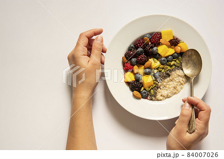 Healthy breakfast with oatmeal porridge, fruits and nuts. Blueberry, mango, pumpkin seeds, nuts, coffee cup. Top view white background. Female hands holding the plate. 84007026