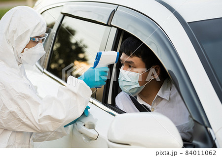 doctor in protective PPE suit using infrared thermometer measuring temperature of man at drive thru station 84011062
