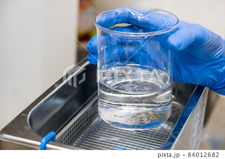 Scientist in blue rubber puts a beaker for dissolving or cleaning into an ultrasound bath. Clinical, toxicological and forensic analysis. Scientist in blue rubber puts a beaker for dissolving or cleaning into an ultrasound bath. Clinical, toxicological and forensic analysis. 84012682