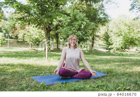 a cheerful woman with a smile on her face is doing yoga sitting in the park in the lotus position 84016670