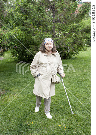 Sad elderly woman with a cane in a jacket walking along the green grass in the park Sad elderly woman with a cane in a jacket walking along the green grass in the park 84019218