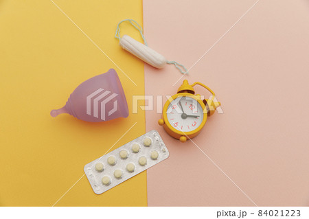 Top view of sanitary cup with pain relief pills, tampon and an alarm clock on colorful background. Menstruation cycle period concept 84021223