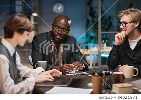 Group of business people sitting at the table discussing and using laptop at work during their meeting till late evening at office 84021871