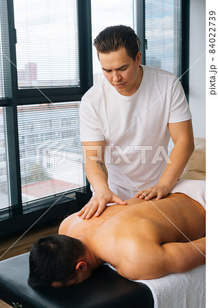 Vertical shot of professional male masseur with strong tattooed hands massaging back and shoulder of muscular sports man lying on stomach Vertical shot of professional male masseur with strong tattooed hands massaging back and shoulder of muscular sports man lying on stomach 84022739