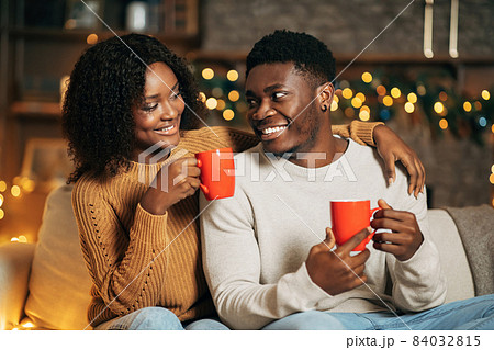 Joyful black couple hugging and drinking coffee, looking at each other, sitting on couch in decorated room on Christmas 84032815