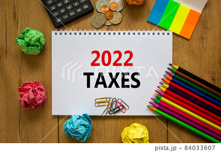 2022 taxes new year symbol. White note with words 2022 taxes on beautiful wooden table, colored paper, colored pencils, paper clips, coins and calculator. Business and 2022 taxes new year concept. 84033607