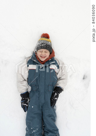 Grimacing boy in a warm jumpsuit and hat lies in the snow. Winter holidays. Child is playing in the snow. Top view 84036090