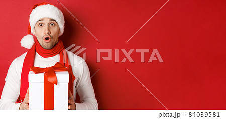 Concept of winter holidays, christmas and lifestyle. Close-up of surprised handsome guy in santa hat and scarf, looking amazed and holding new year gift, standing over red background Concept of winter holidays, christmas and lifestyle. Close-up of surprised handsome guy in santa hat and scarf, looking amazed and holding new year gift, standing over red background 84039581